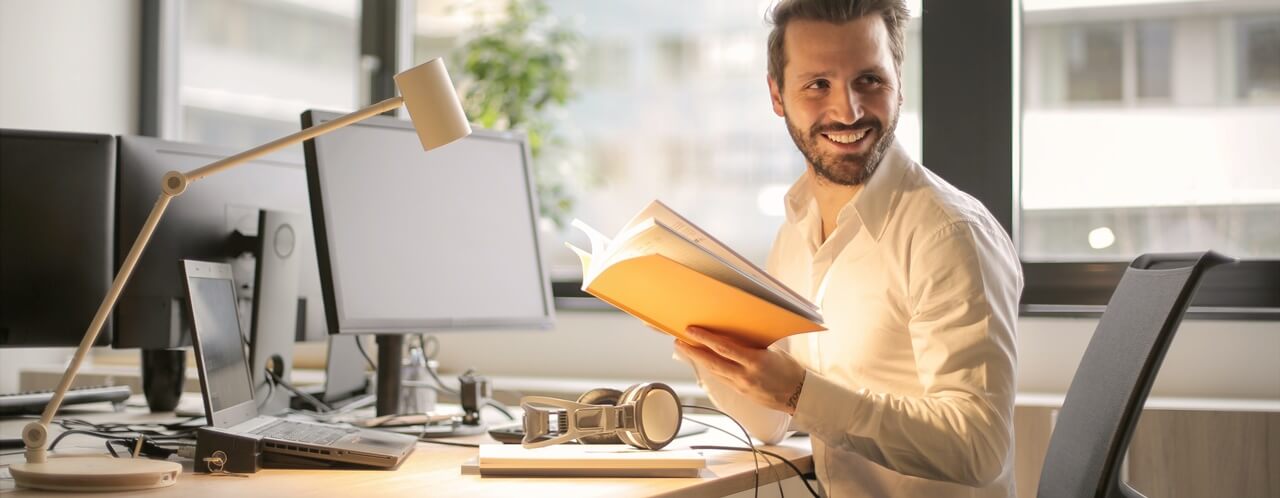A happy man on his computer while holding a book.